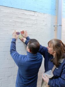 Two people holding ceramic tiles up to a plain brick wall